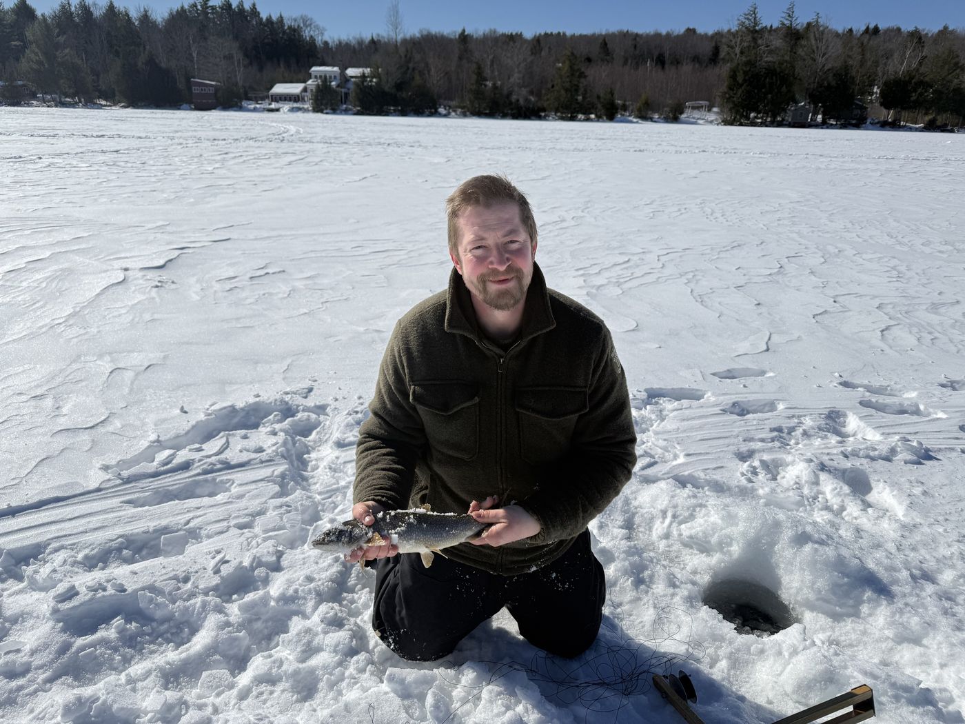 Angler kneeling beside an auger hole with a chain pickerel in Maine