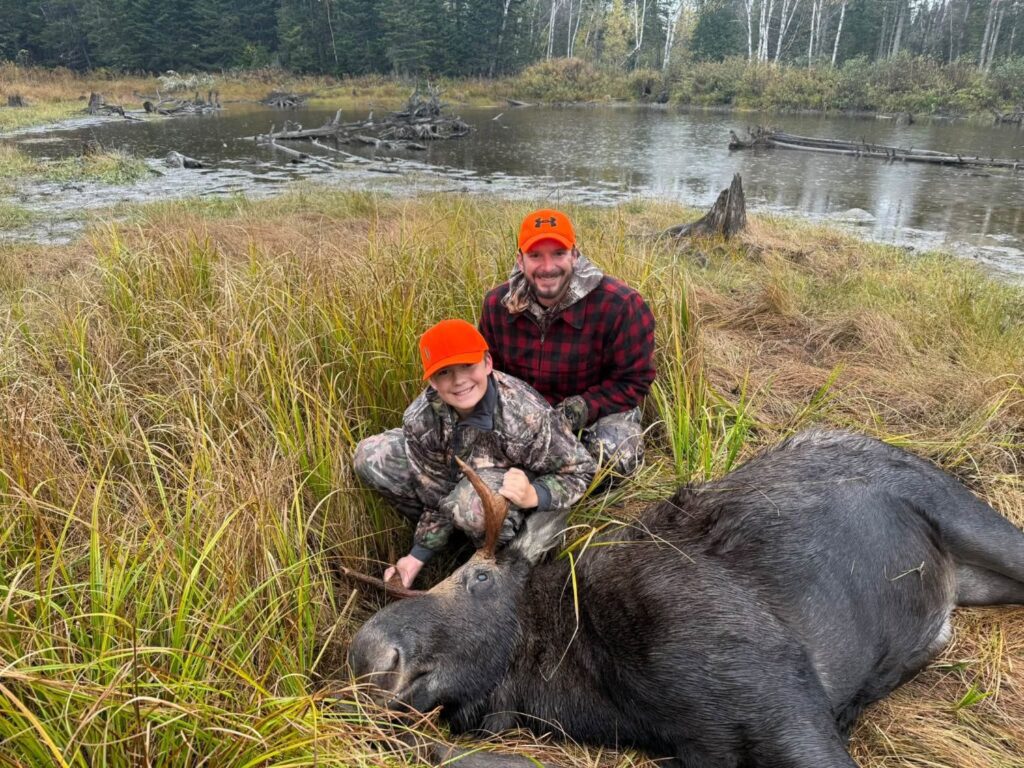 Young hunter and his father kneeling beside a harvested Maine bull moose beside a beaver flowage