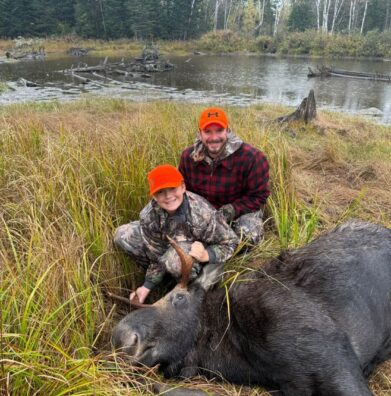 Young hunter and his father kneeling beside a harvested Maine bull moose beside a beaver flowage